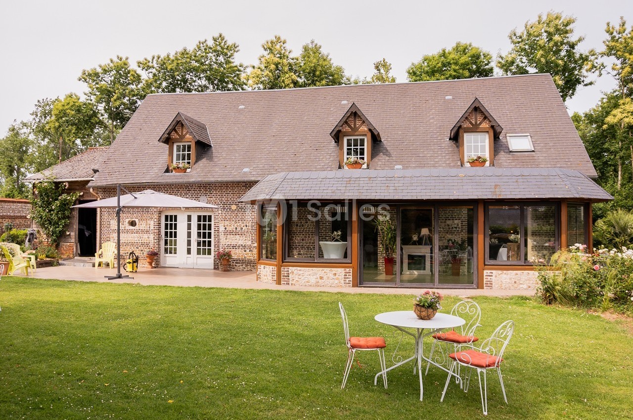 Maison en briques avec toit en ardoise, baies vitrées, terrasse aménagée et jardin avec table et chaises blanches.