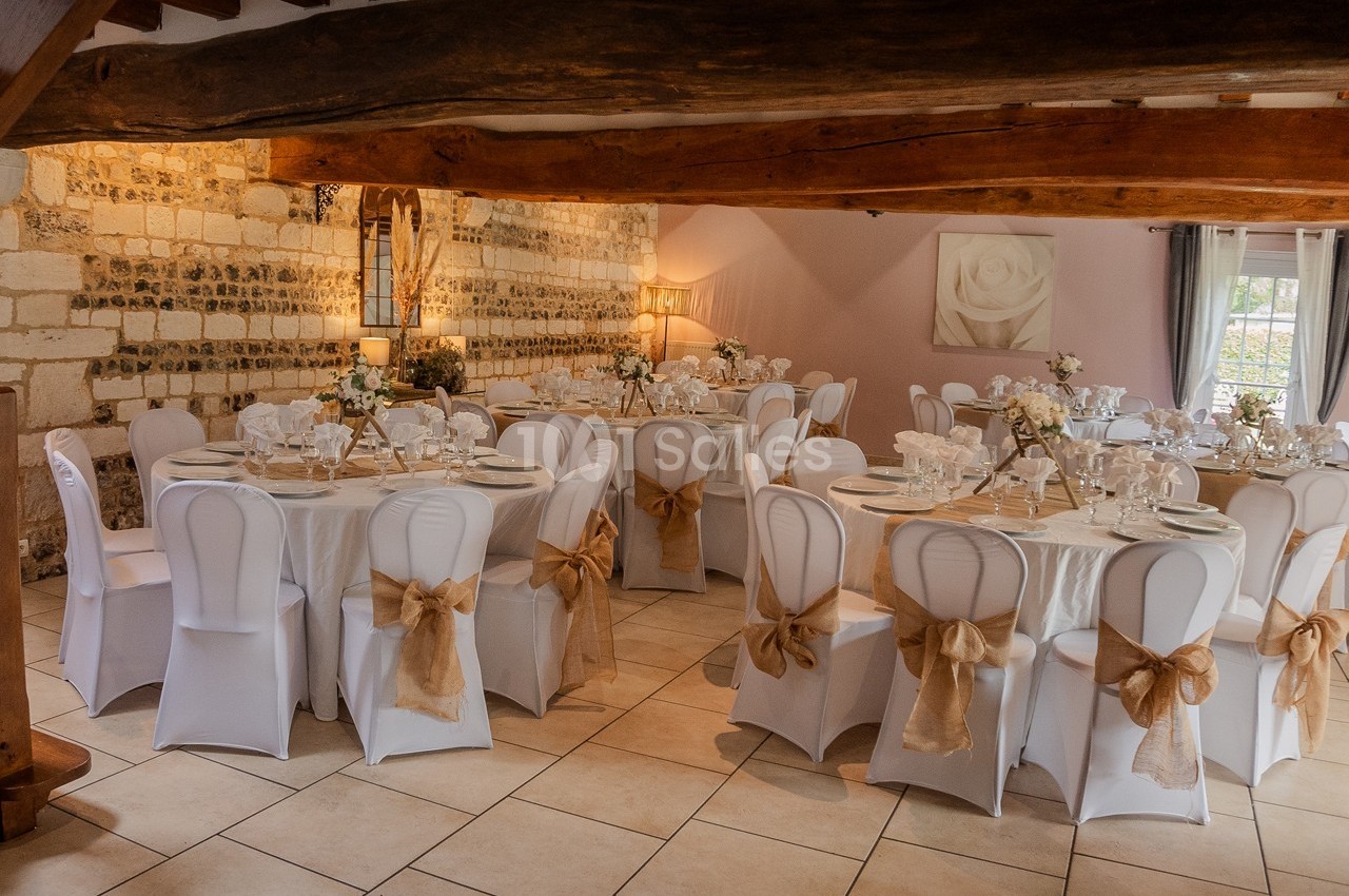 Salle de réception décorée avec des tables rondes, nappes blanches, chaises ornées de nœuds beige et poutres apparentes.