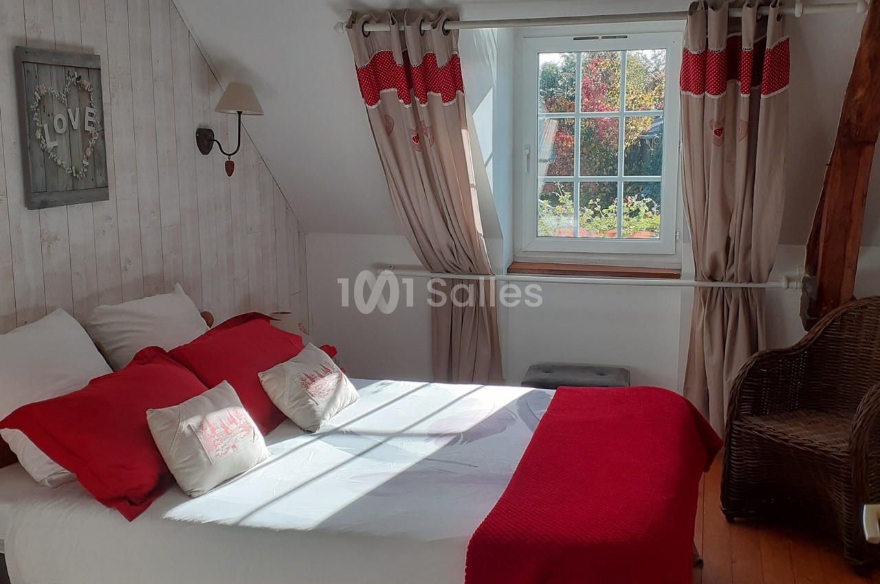 Chambre lumineuse avec lit double, draps rouges et blancs, fenêtre donnant sur un jardin, fauteuil en osier à droite.