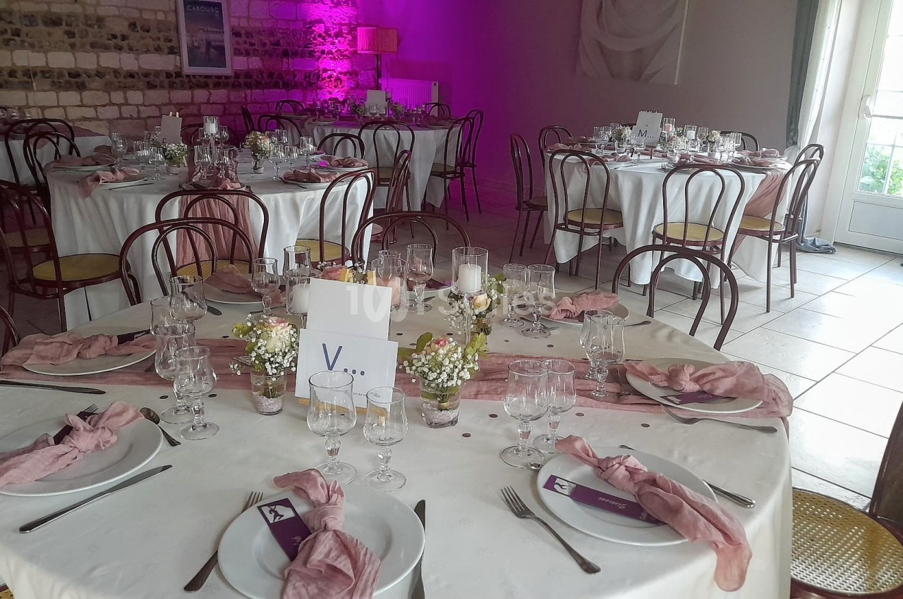 Salle de réception décorée avec des tables rondes, nappes blanches, serviettes roses et centres de table fleuris.