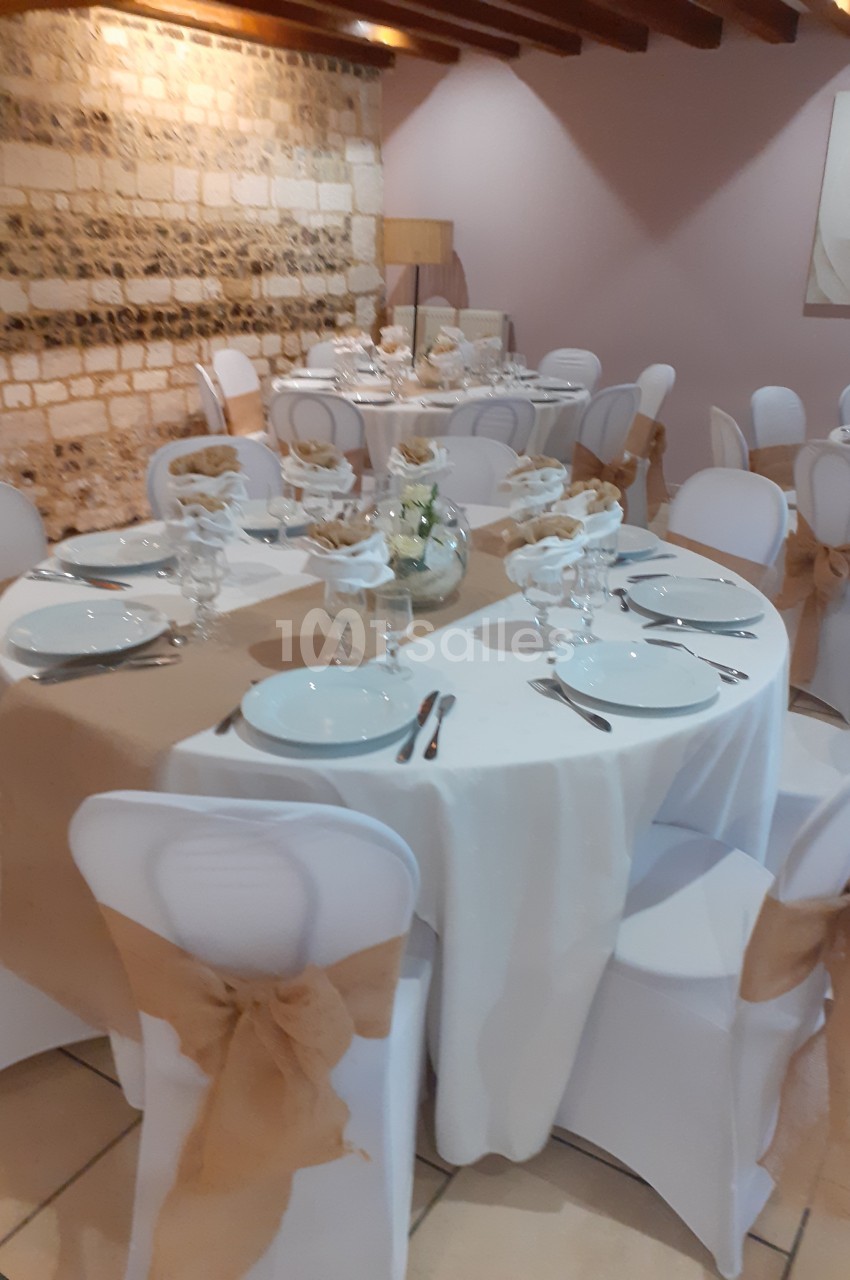 Salle décorée pour un événement avec des tables rondes dressées, nappes blanches et chaises ornées de nœuds en toile de jute.