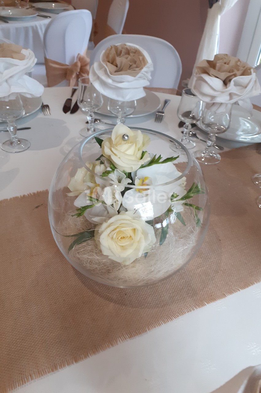 Centre de table avec roses blanches et fleurs dans un vase en verre rond, sur une nappe en toile de jute.