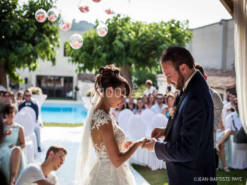 Un couple échange des alliances lors d'une cérémonie de mariage en extérieur, entouré d'invités assis.