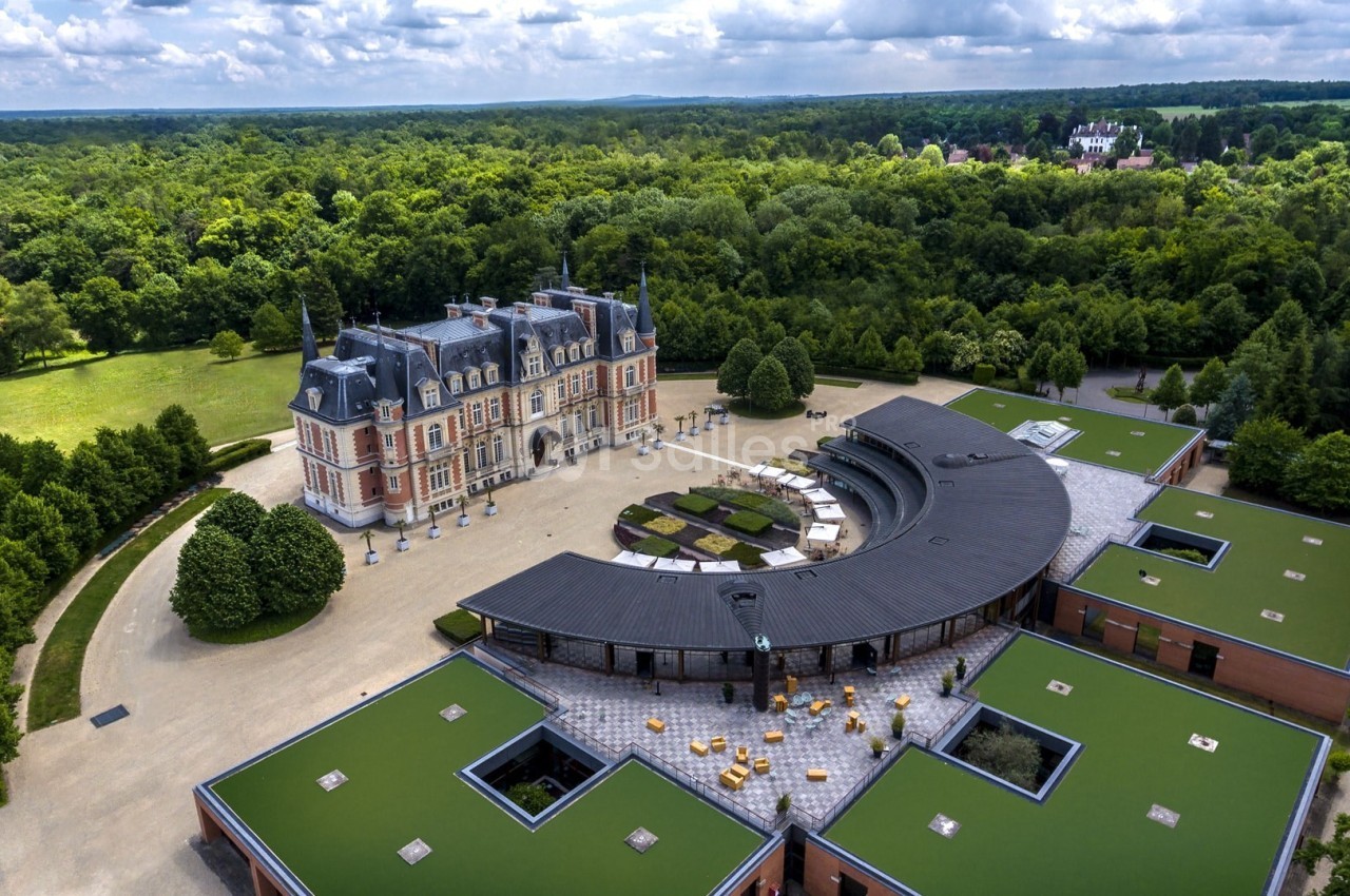 Vue aérienne d'un château entouré de forêts, avec des bâtiments modernes et des toits végétalisés au premier plan.