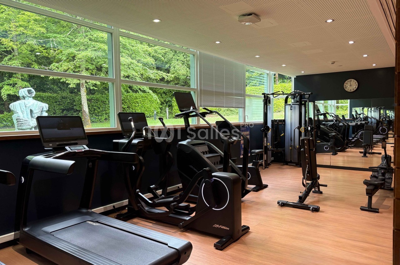 Salle de sport lumineuse avec tapis de course, vélos elliptiques, machines de musculation et miroir mural.