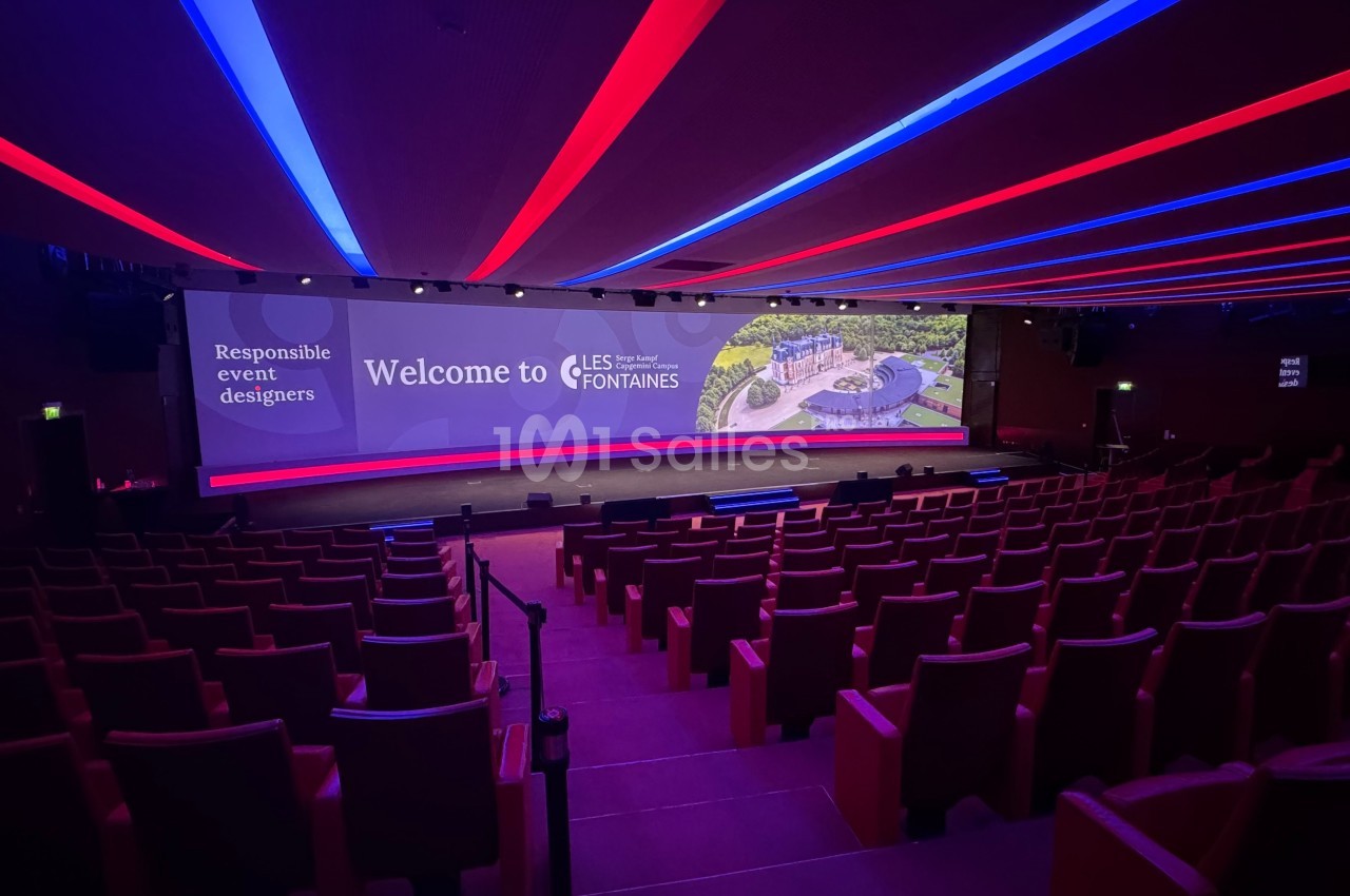 Salle de conférence vide avec sièges rouges, grand écran affichant ’Welcome to Les Fontaines’ et éclairage coloré au plafond.