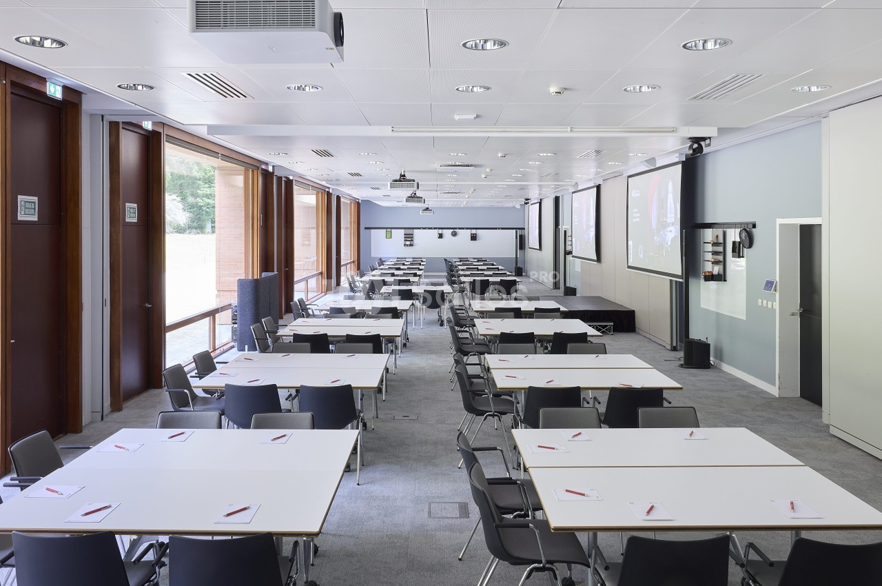 Salle de conférence lumineuse avec des tables blanches, des chaises noires et des équipements audiovisuels.