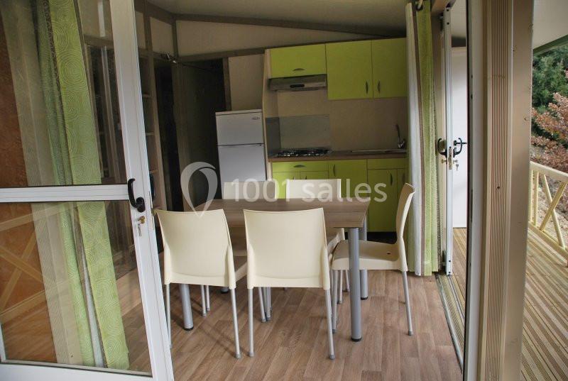 Salle à manger avec table et chaises blanches, ouverte sur une cuisine équipée et une terrasse en bois.