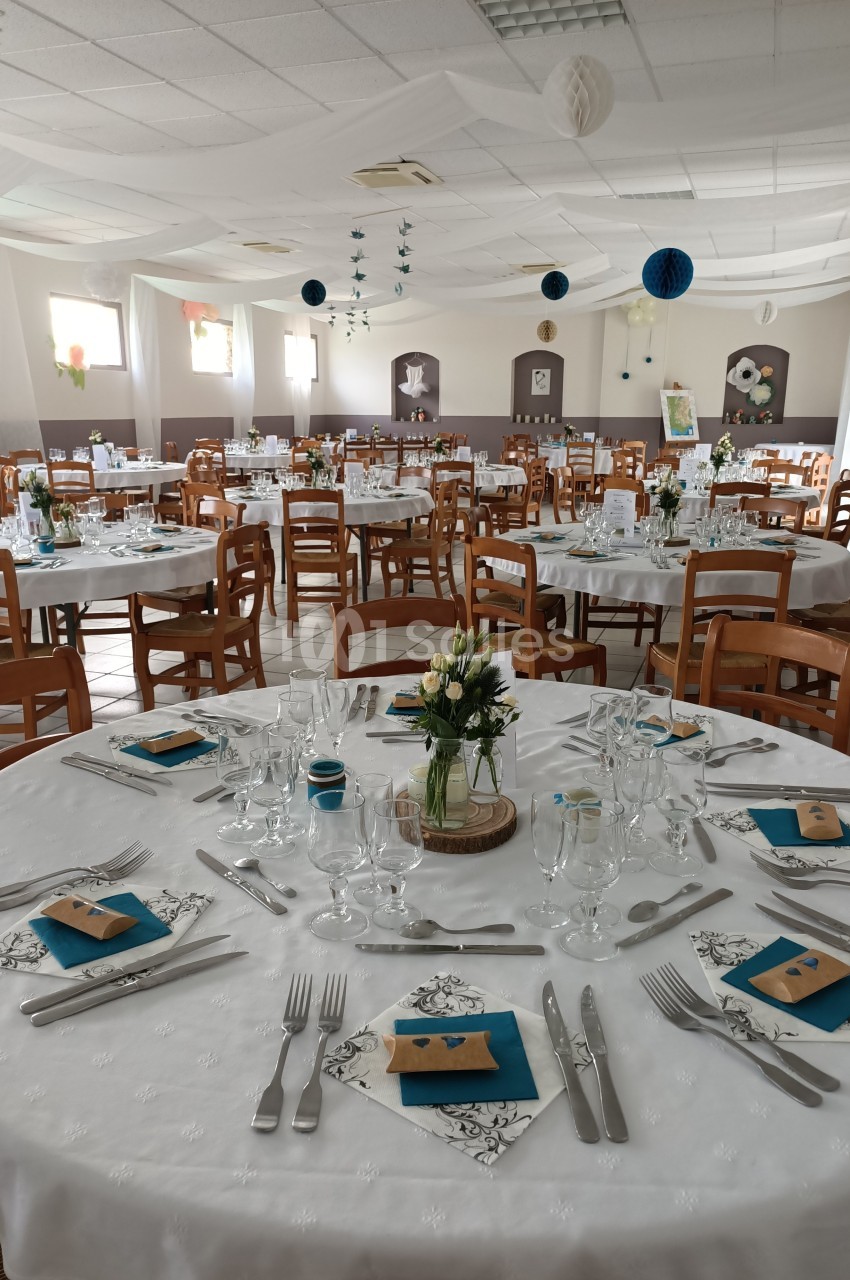 Salle de réception décorée avec des tables rondes dressées, nappes blanches, serviettes bleues et centres de table floraux.