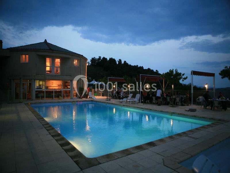 Grande piscine éclairée en soirée devant une maison, avec des personnes rassemblées sur une terrasse adjacente.