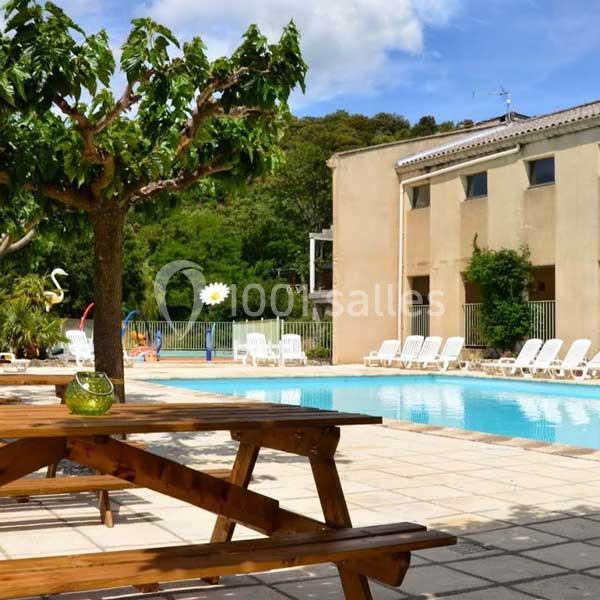 Piscine extérieure entourée de transats, avec une table en bois au premier plan et des arbres en arrière-plan.