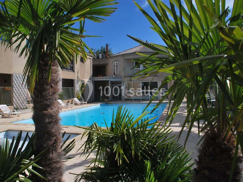 Piscine extérieure entourée de palmiers avec des transats et un bâtiment en arrière-plan sous un ciel bleu.