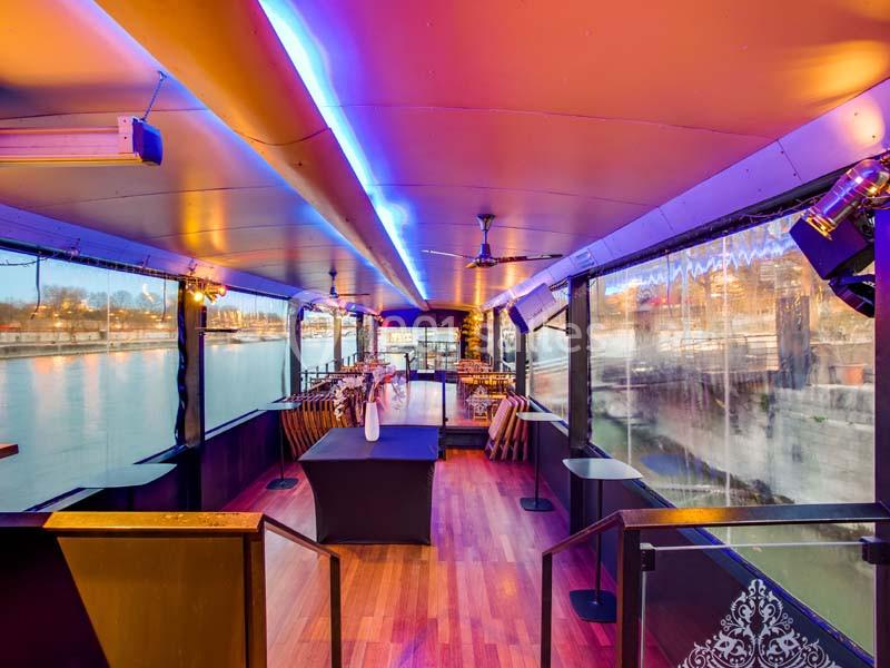 Intérieur d'une péniche aménagée avec parquet, tables dressées et éclairage coloré, vue sur l'eau à travers les baies…
