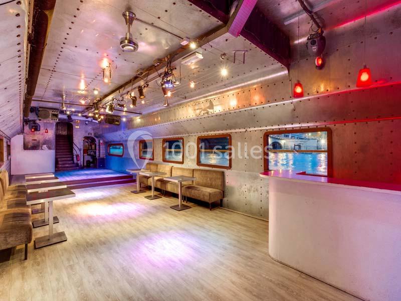 Intérieur d'un espace lumineux avec murs métalliques, tables, banquettes et vue sur l'eau à travers des hublots.