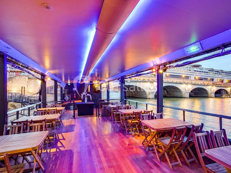 Terrasse couverte avec tables en bois, éclairage coloré et vue sur un pont au-dessus d'une rivière.
