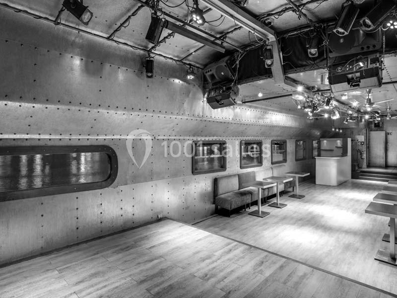 Salle intérieure au design industriel avec murs métalliques rivetés, tables, banquettes et éclairage suspendu.
