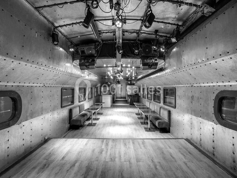 Intérieur d'un wagon métallique aménagé avec des banquettes, un sol en bois et un éclairage suspendu.