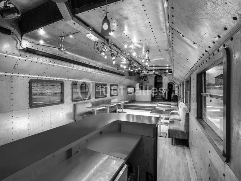 Intérieur d'un wagon aménagé en espace public avec des tables, des banquettes et un éclairage suspendu.