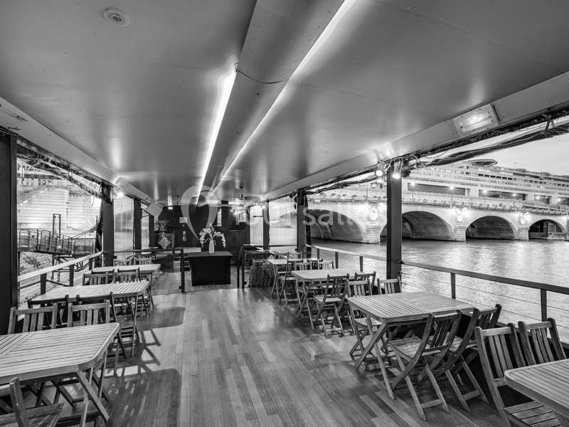 Salle avec tables et chaises en bois sur une péniche, offrant une vue sur un pont et une rivière en arrière-plan.