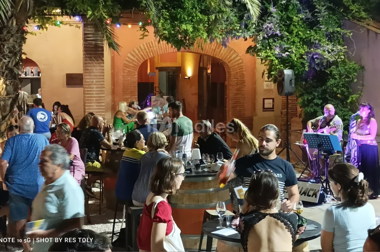 Groupe de personnes attablées en soirée dans une cour extérieure éclairée, avec des musiciens jouant en arrière-plan.