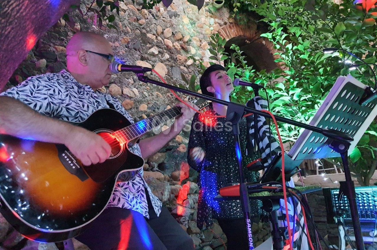 Deux musiciens jouent de la guitare et chantent en extérieur, entourés de végétation et éclairés par des lumières colorées.