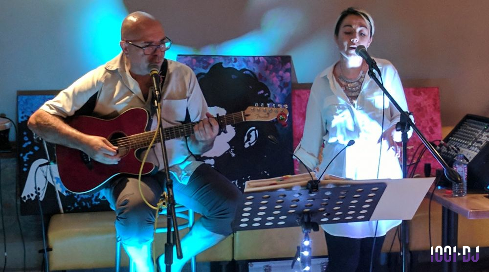 Un duo chante et joue de la guitare sur une scène éclairée, avec des tableaux en arrière-plan.