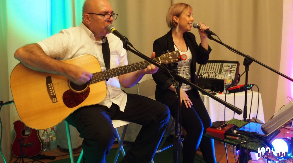 Un homme joue de la guitare acoustique et une femme chante, tous deux assis devant des micros dans un cadre lumineux.