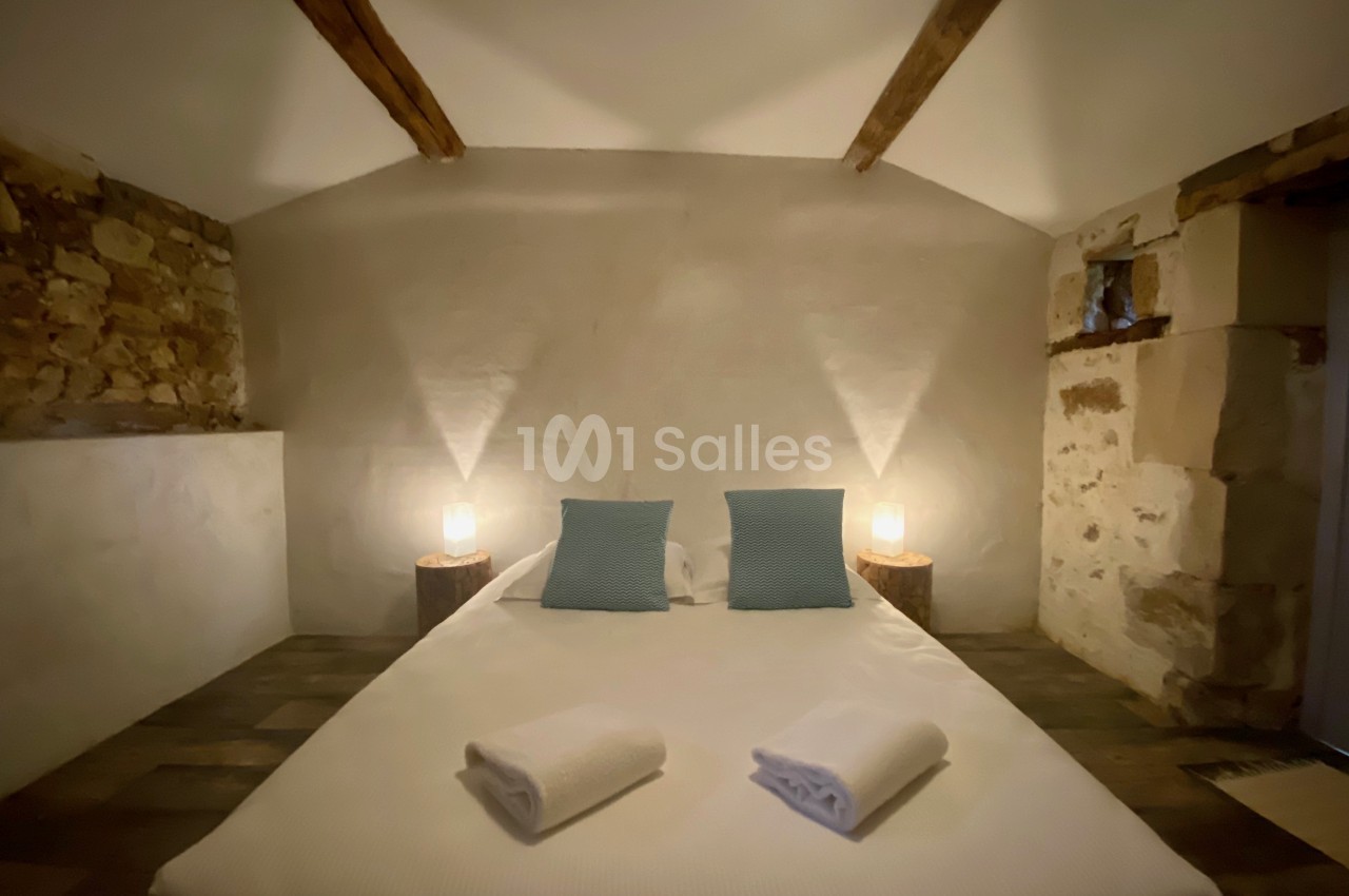 Lit double avec draps blancs, deux coussins bleus, éclairage doux et murs en pierre dans une chambre rustique.