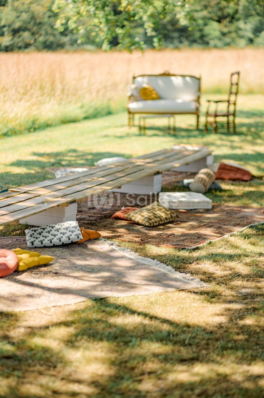 Banc en bois avec coussins colorés sur des tapis en plein air, entouré d'herbe et de mobilier vintage.
