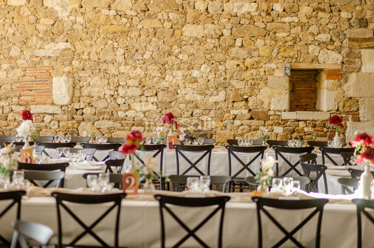 Salle décorée pour un événement avec des tables alignées, nappes blanches, fleurs rouges et murs en pierre apparente.