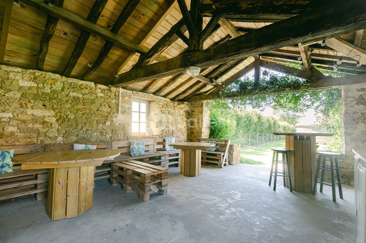 Terrasse couverte en pierre et bois avec mobilier rustique, ouverte sur un jardin verdoyant.
