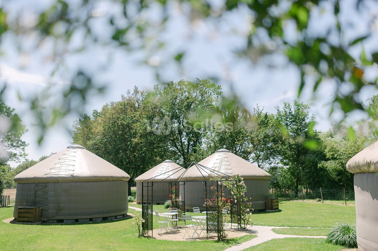 Trois yourtes beige clair entourées de verdure, avec un salon de jardin sous une pergola au premier plan.