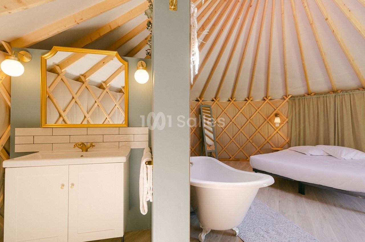 Intérieur d'une yourte avec salle de bain équipée d'une baignoire, lavabo et miroir doré, et un lit en arrière-plan.