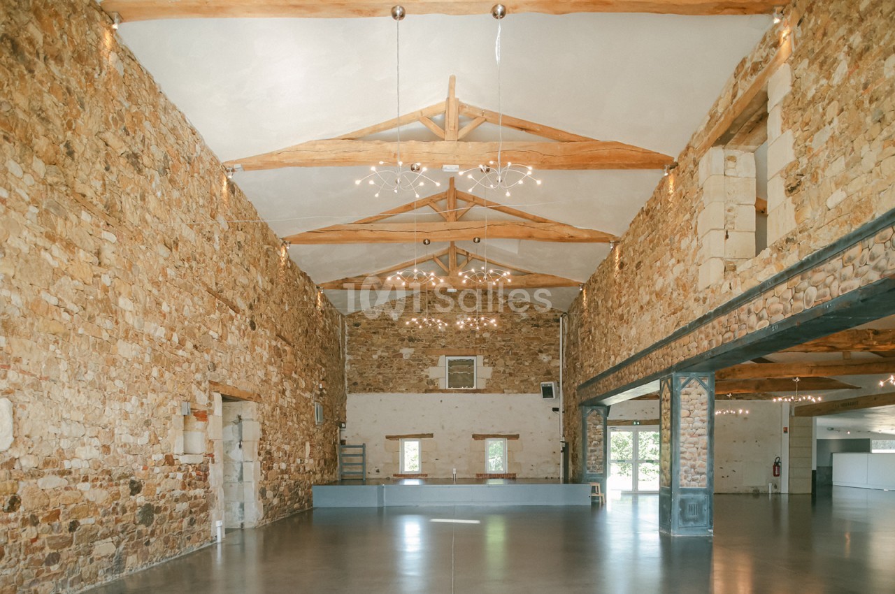 Salle spacieuse avec murs en pierre, poutres apparentes et éclairage suspendu, offrant une ambiance lumineuse et rustique.