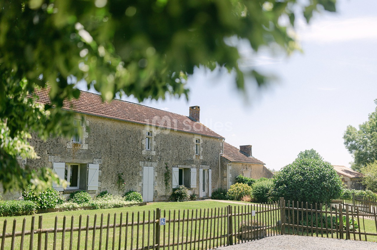 Maison en pierre avec volets blancs entourée de verdure, vue à travers des branches et une clôture en bois.