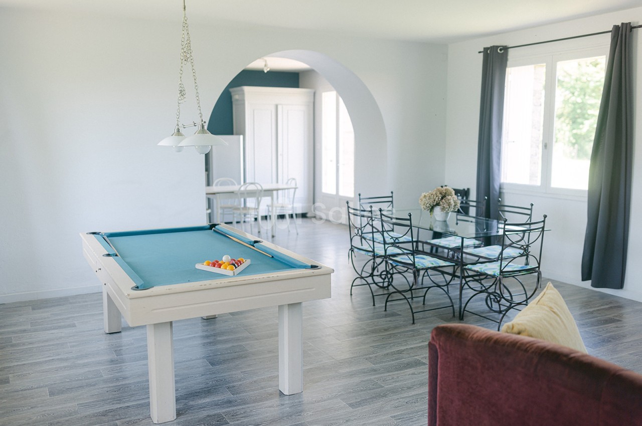 Salle lumineuse avec table de billard, coin repas avec chaises en métal et grande fenêtre donnant sur l'extérieur.