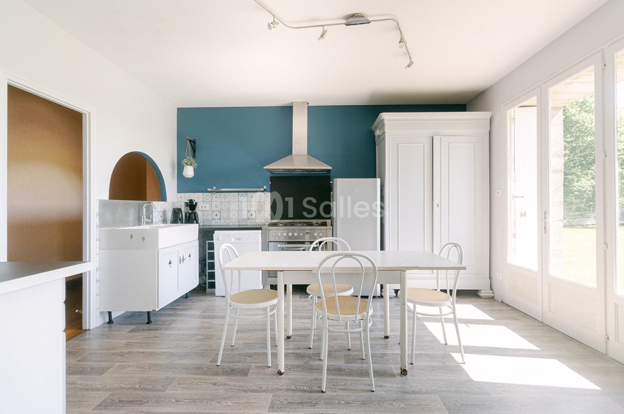 Cuisine lumineuse avec table blanche, chaises en métal, mur bleu, sol en bois clair et grande fenêtre donnant sur l…