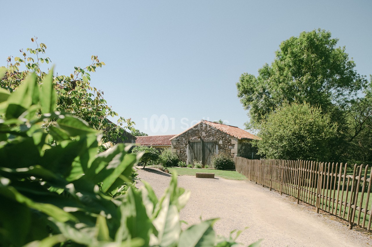 Chemin de gravier menant à une maison en pierre avec toit en tuiles, entourée de verdure et d'une clôture en bois.