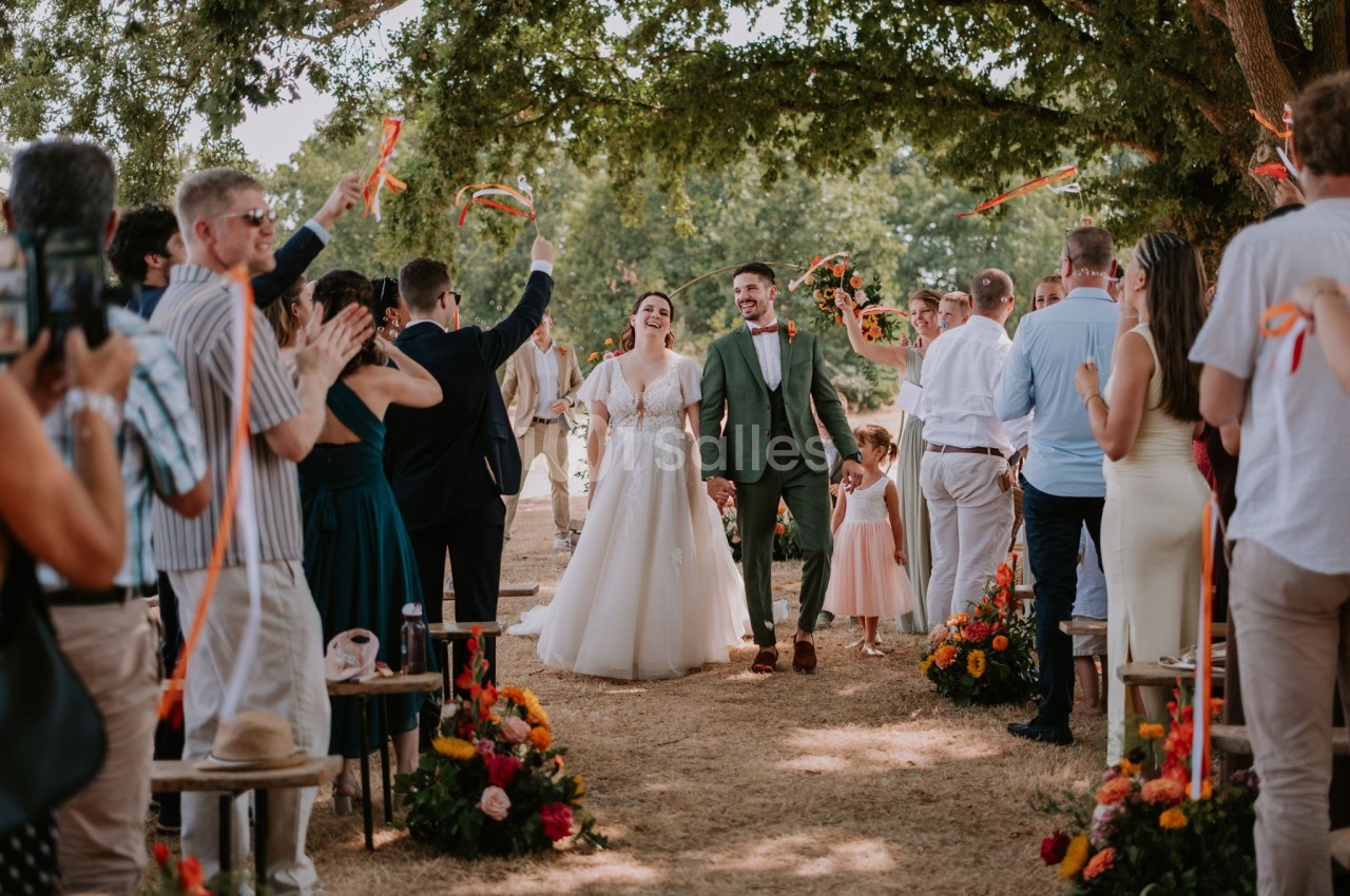 Un couple de mariés souriant marche sous une allée festive entourée d'invités applaudissant en extérieur.