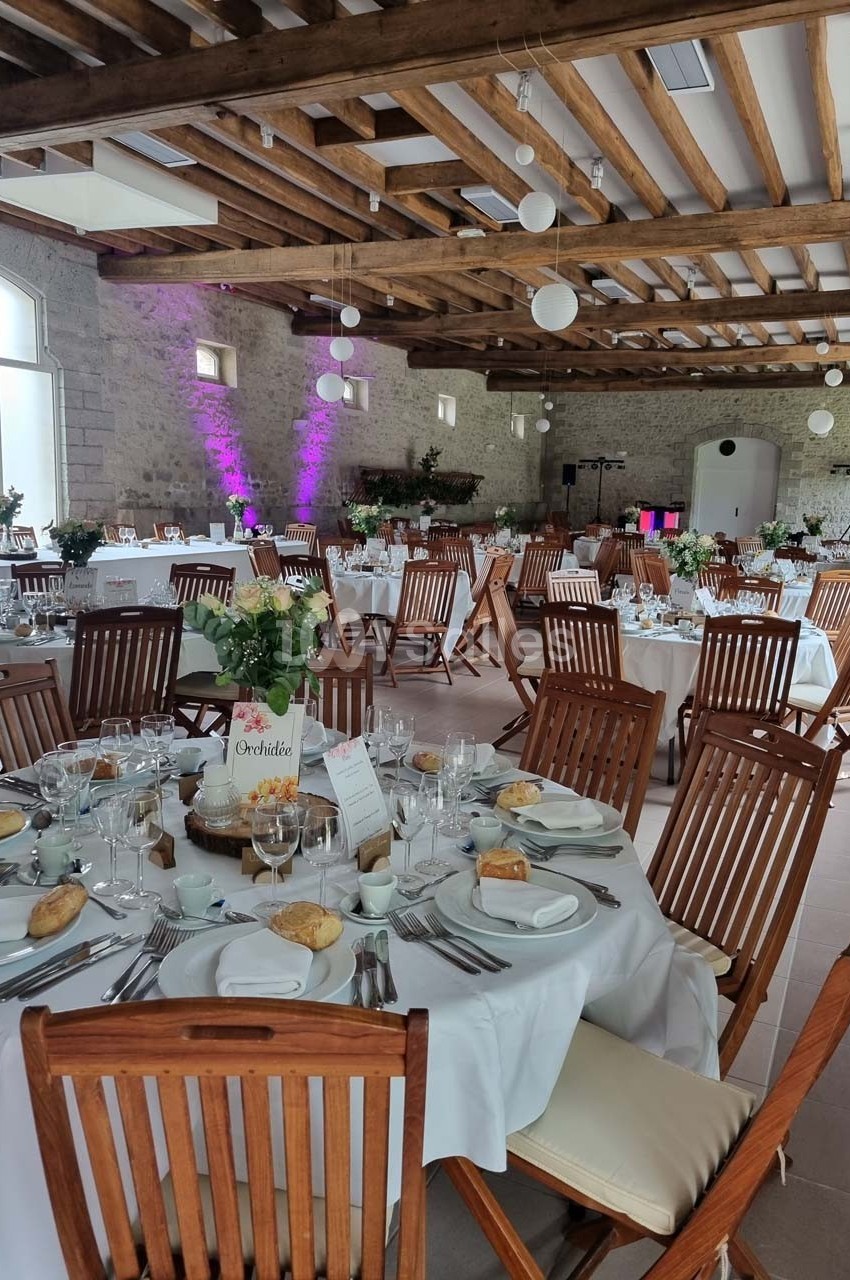 Salle de réception décorée avec des tables rondes dressées, chaises en bois et éclairage tamisé.
