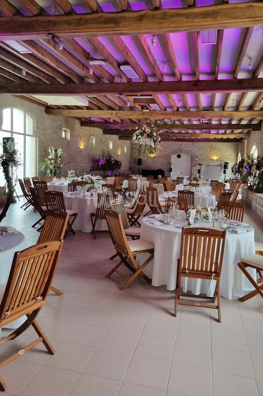 Salle de réception décorée avec des tables rondes, nappes blanches, chaises en bois et éclairage violet.