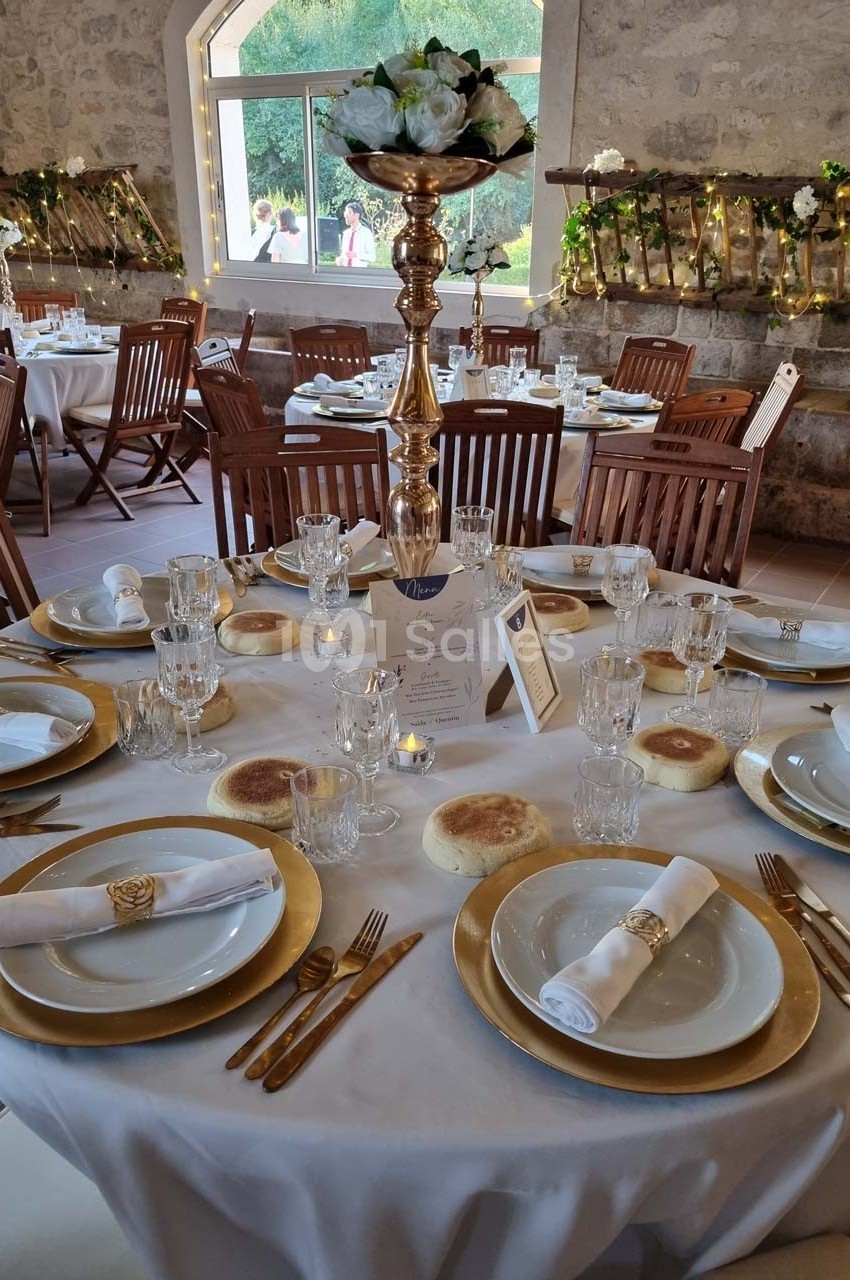 Table de réception élégamment dressée avec vaisselle dorée, serviettes roulées et centre de table fleuri dans une salle en…