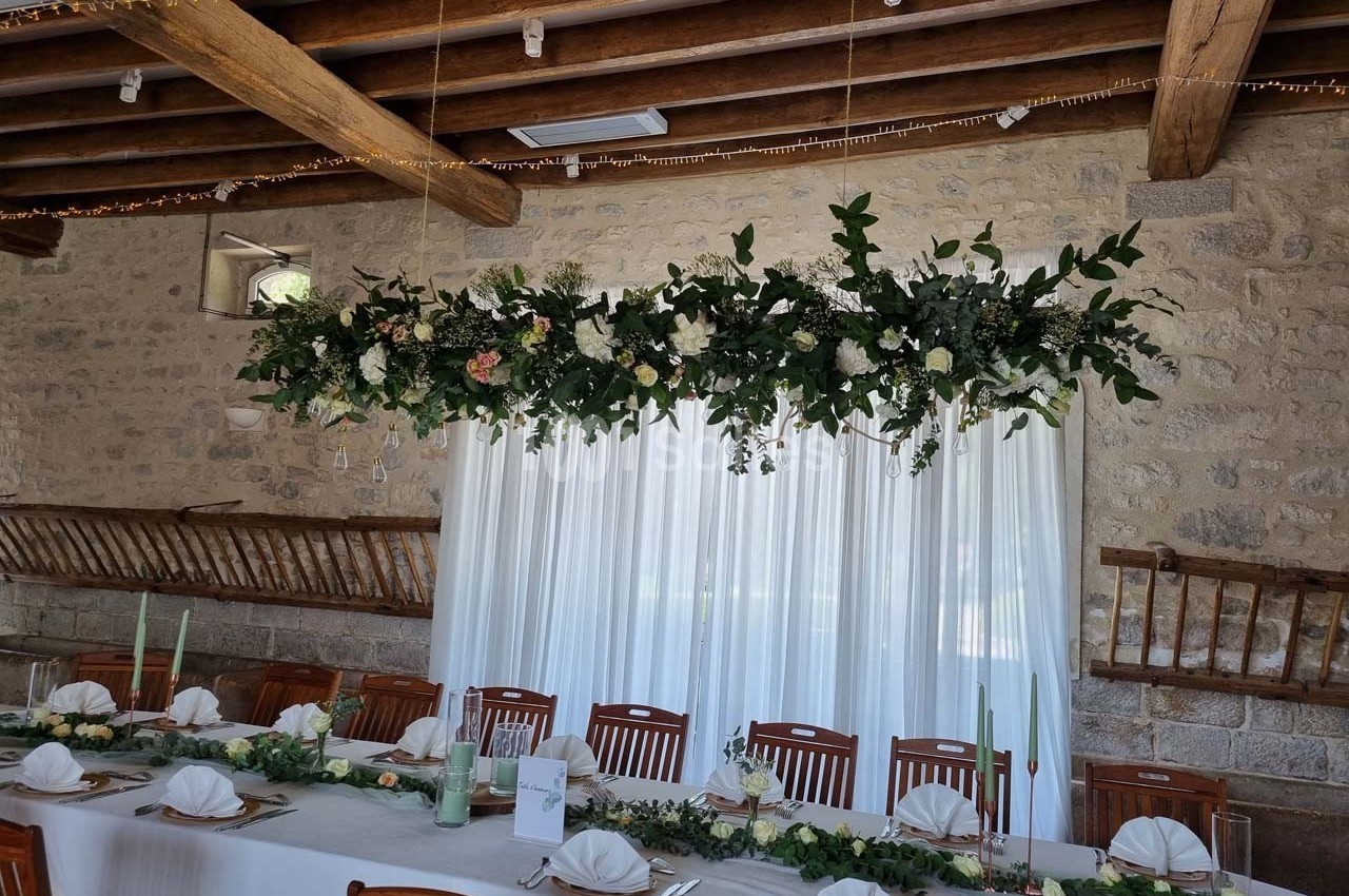 Décoration florale suspendue au-dessus d'une table de réception dressée dans une salle en pierre avec poutres apparentes.