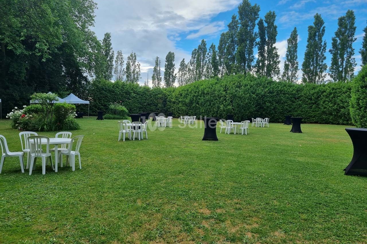 Chaises et tables blanches disposées sur une pelouse avec des mange-debout noirs, entourées de haies et d'arbres.