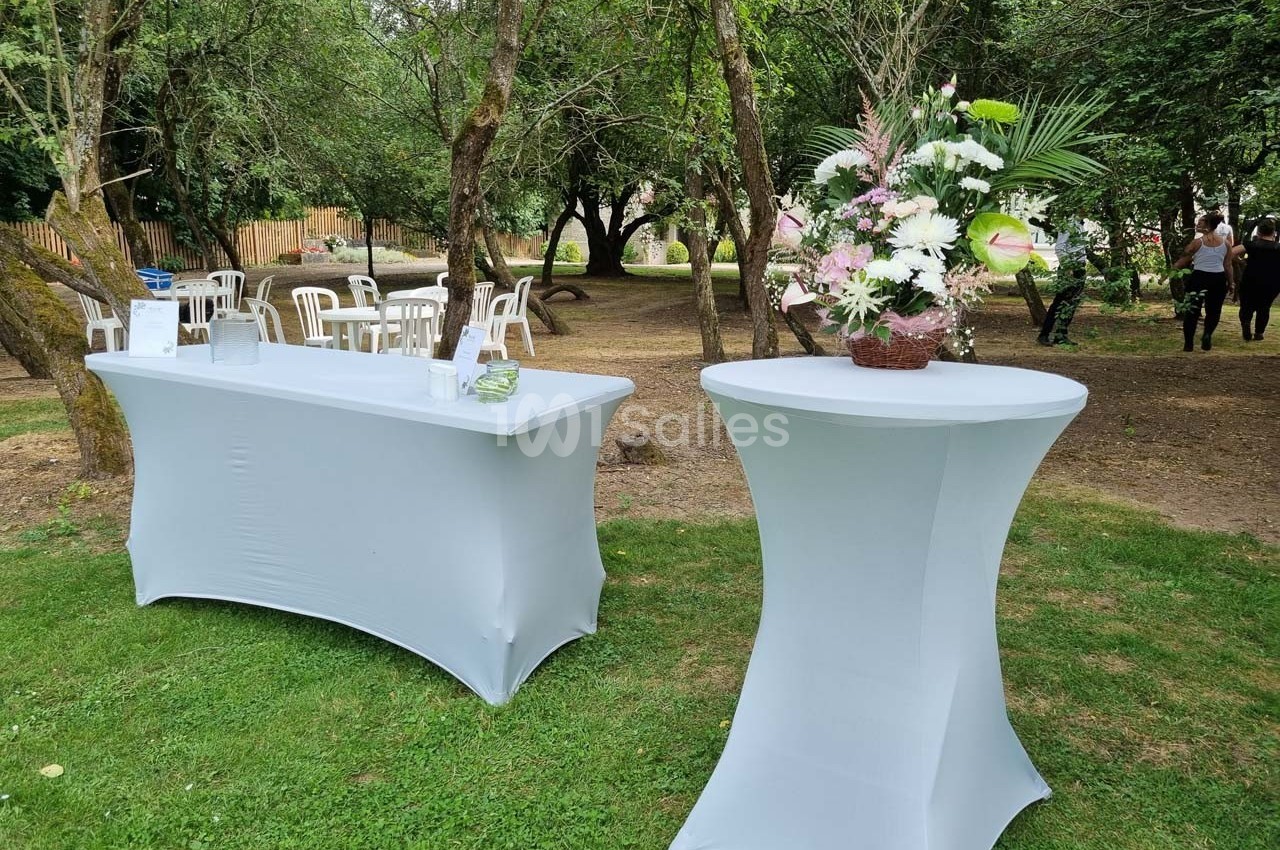Table dressée avec nappe blanche et décor floral, installée en extérieur sur une pelouse entourée d'arbres.