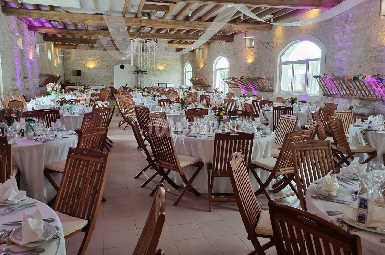Salle de réception lumineuse avec tables rondes dressées, chaises en bois et décorations élégantes.