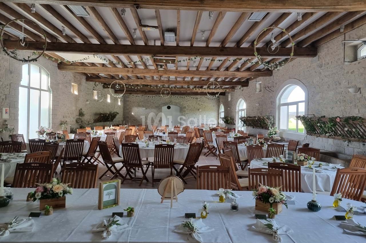 Salle de réception décorée avec des tables dressées, chaises en bois et ornements floraux, sous un plafond en poutres…