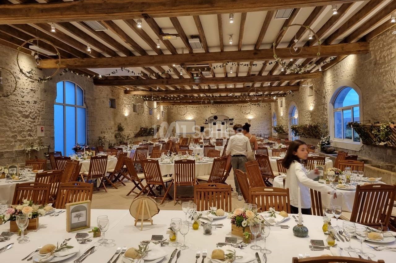 Salle de réception en pierre avec tables dressées pour un événement, décorée de fleurs et éclairée par des lumières…