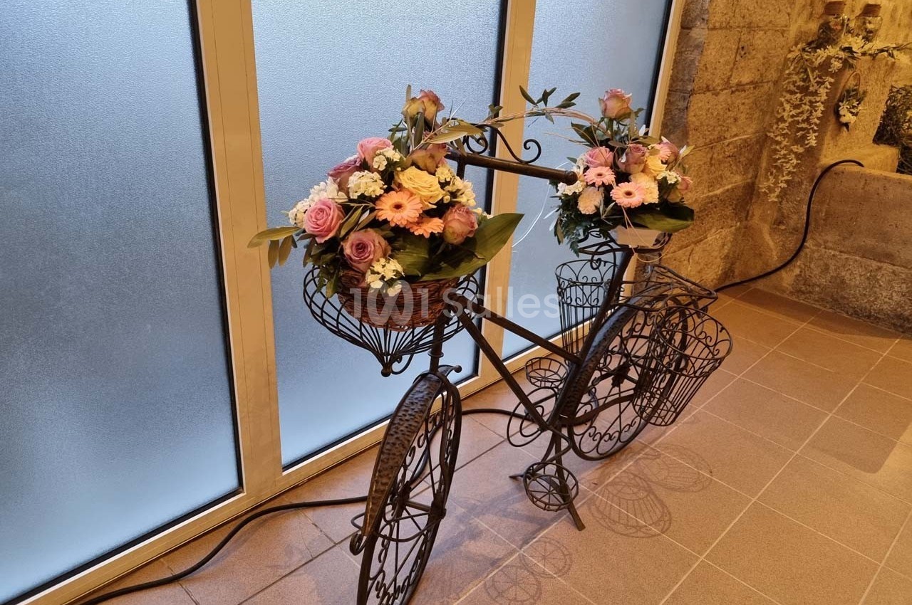 Vélo décoratif en métal noir avec des paniers garnis de fleurs roses et blanches, placé devant une fenêtre givrée.