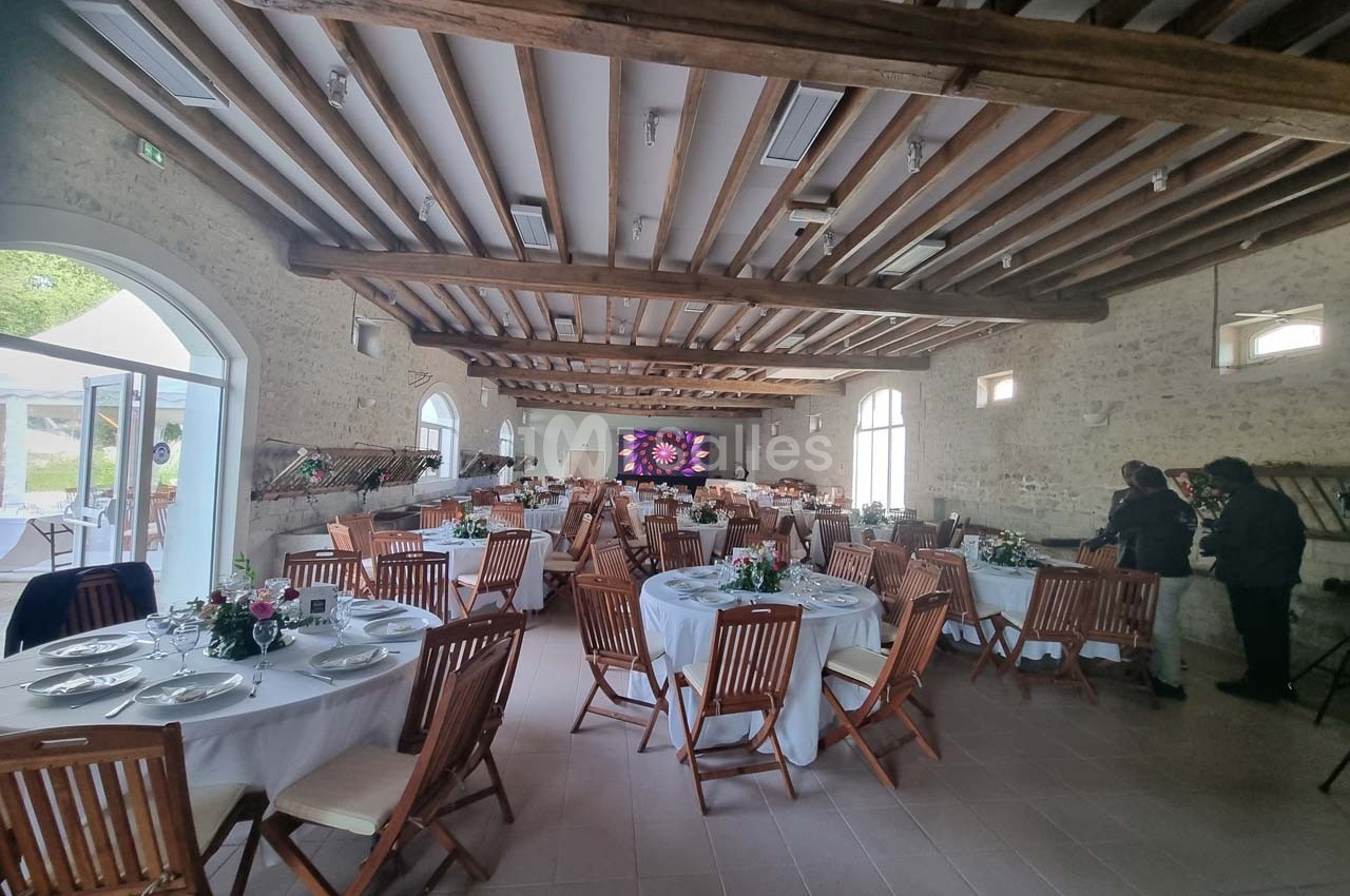 Salle de réception avec tables rondes dressées, chaises en bois, poutres apparentes et écran au fond.